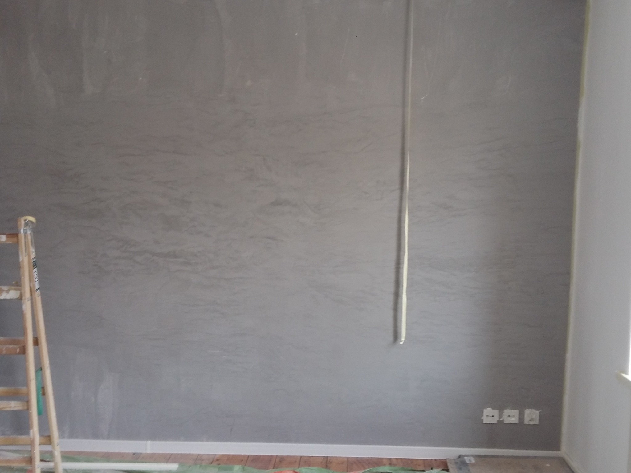 Ściana w szarym odcieniu z efektem dekoracyjnym, widoczna drabina malarska, zabezpieczona podłoga i wystające kable elektryczne.
