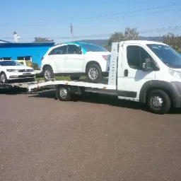 Biała laweta z dwoma białymi samochodami: SUV na platformie i sedan na przyczepie, na tle błękitnego nieba i zabudowań.