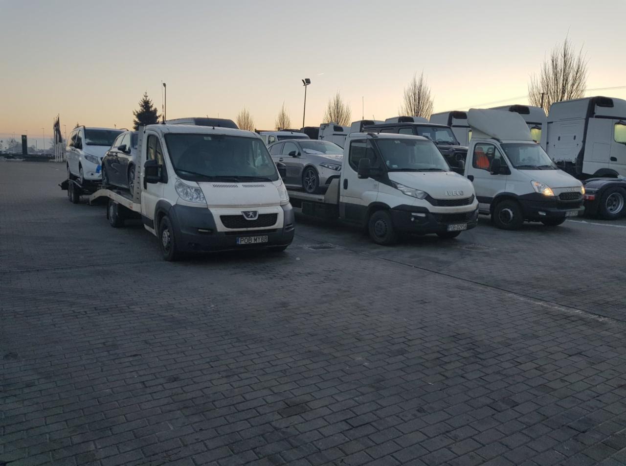 Kilka lawet na parkingu, jedna Peugeot z lawetą przewozi dwa samochody osobowe, obok Iveco z podobnym ładunkiem, w tle inne pojazdy i drzewa.