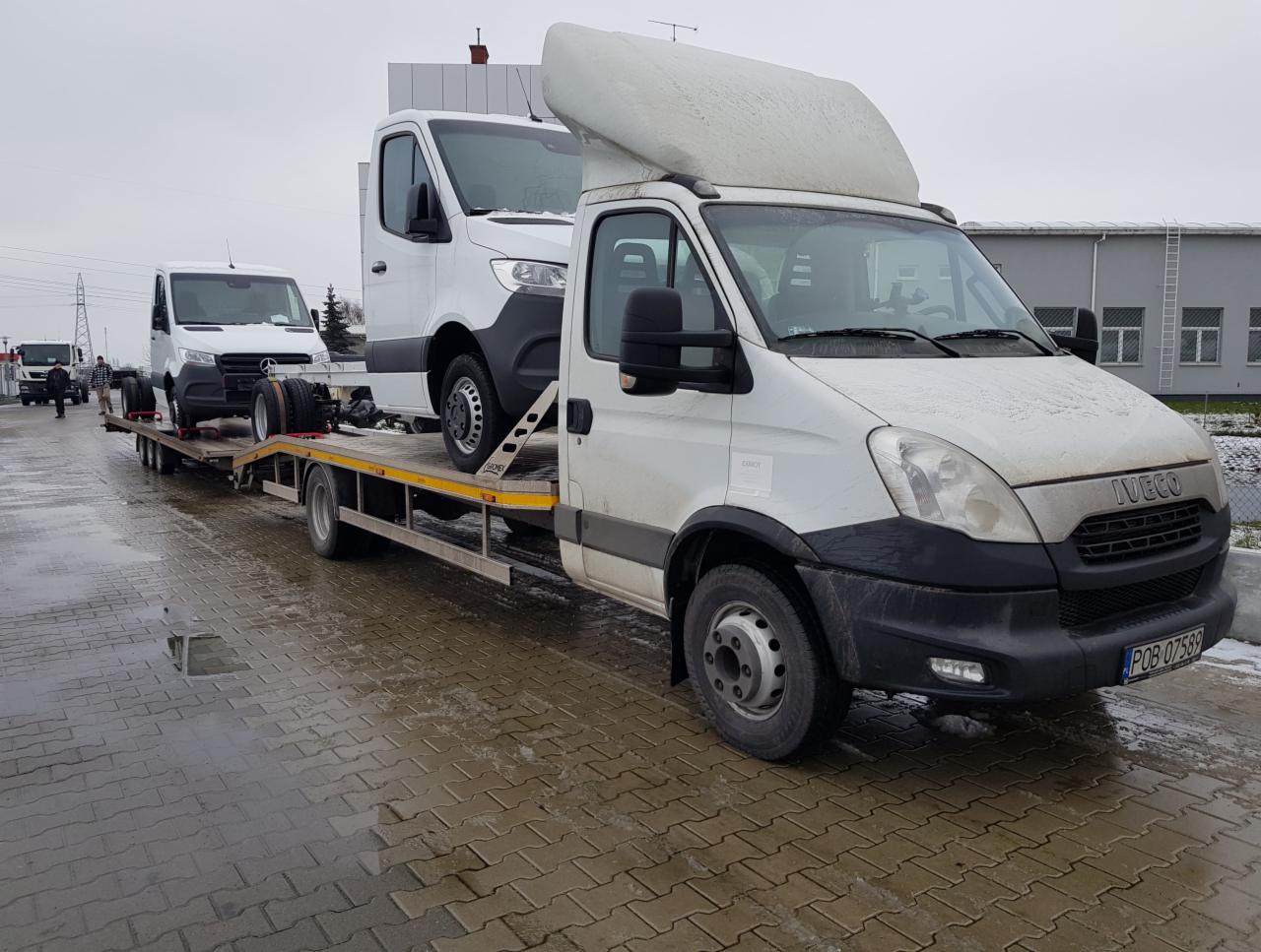 Biała laweta Iveco z dwoma białymi samochodami dostawczymi na platformie, zaparkowana na mokrym bruku w pochmurny dzień.