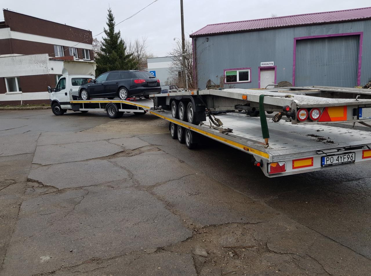 Czarny samochód kombi zabezpieczony pasami na platformie lawety, z widoczną tablicą rejestracyjną PO 4YF96, zaparkowanej na popękanym asfalcie, z budynkiem magazynowym w tle.