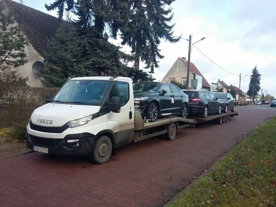 Biała laweta marki Iveco przewozi trzy czarne samochody osobowe na platformie w Obornikach, widok z boku na tle domów i drzew.