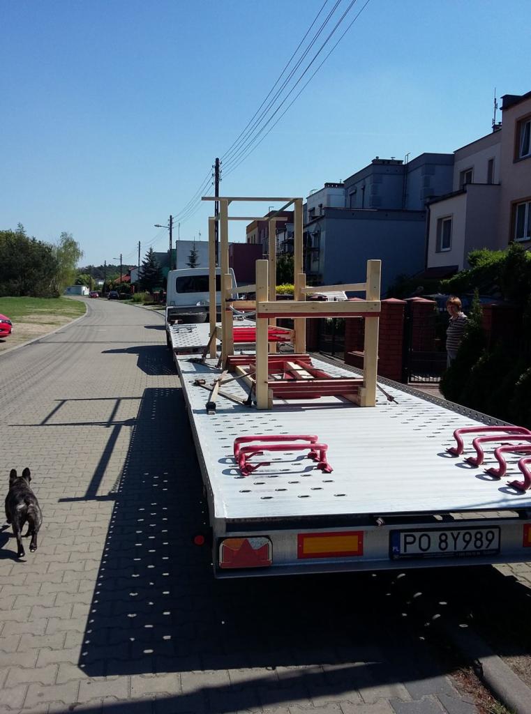 Laweta z drewnianą konstrukcją na platformie, zabezpieczoną czerwonymi uchwytami, zaparkowana na brukowanej ulicy w słoneczny dzień; z tyłu widoczny budynek mieszkalny i osoba.