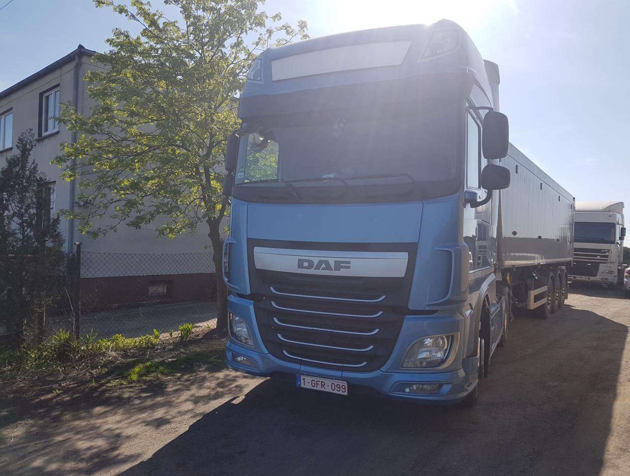 Błękitny ciągnik siodłowy marki DAF z naczepą na żwirowej nawierzchni, z budynkiem mieszkalnym i drzewem w tle, widoczne tablice rejestracyjne.