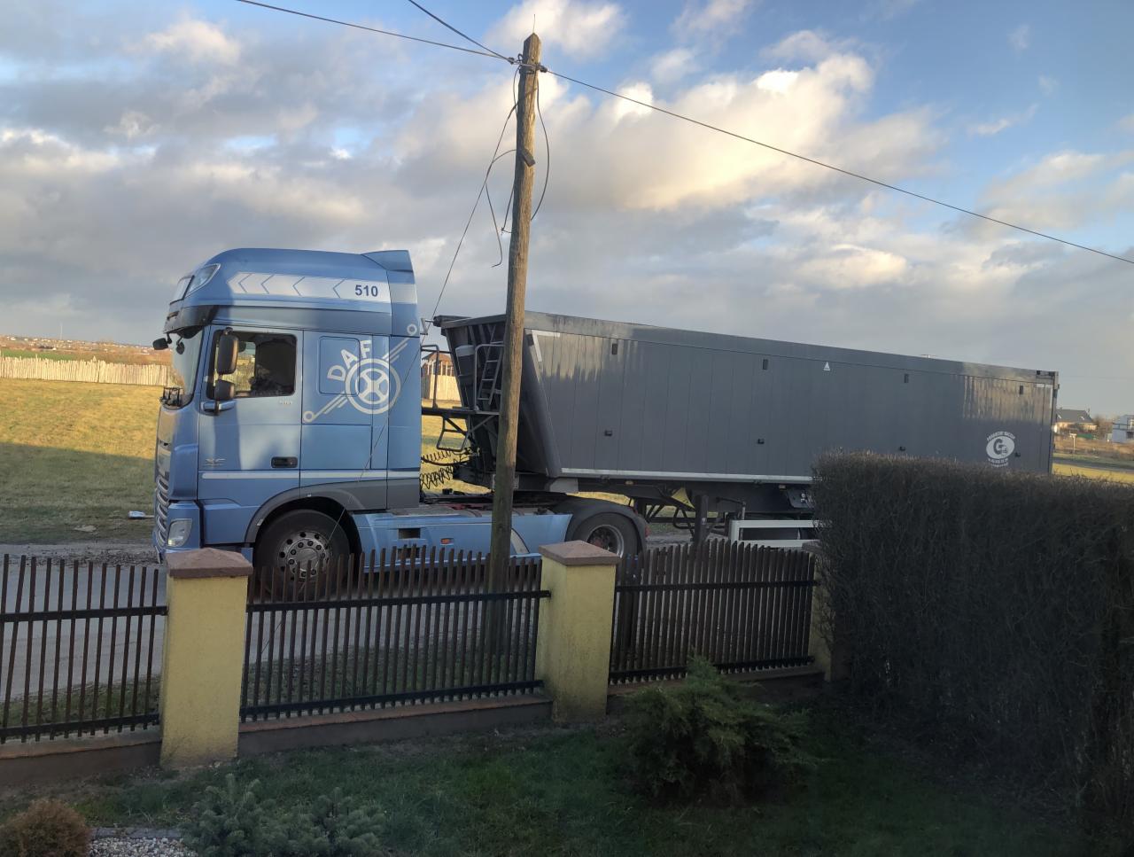 Błękitny ciągnik siodłowy DAF z szarą naczepą typu wywrotka, zaparkowany za niskim płotem w słoneczny dzień.