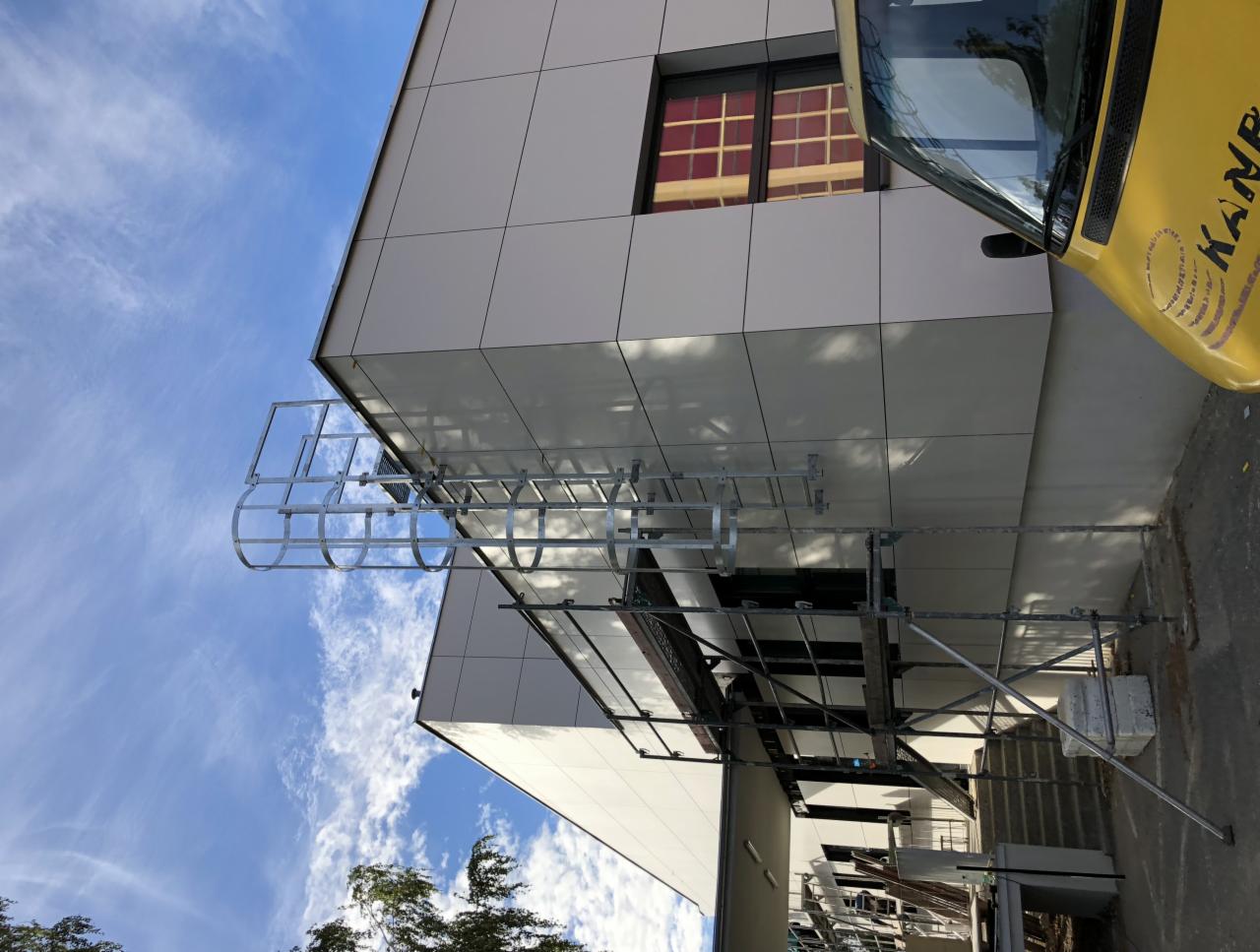 Metalowa drabina ewakuacyjna z koszem ochronnym przymocowana do ściany budynku z jasnymi panelami, widok od dołu, obok zaparkowany żółty samochód dostawczy z logo firmy.