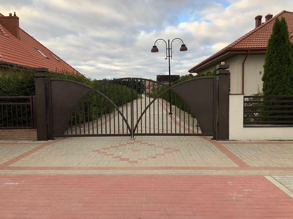 Automatyczna brama wjazdowa z elementami łukowymi, brązowa, widok z zewnątrz na posesję z kostki brukowej i lampą uliczną.