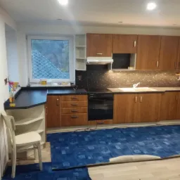 Kuchnia z brązowymi szafkami i czarnym blatem, widoczne niedokończone prace podłogowe z rozłożoną niebieską wykładziną i krzesłem z boku.