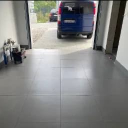 Szare płytki podłogowe w garażu, widok na zewnątrz z zaparkowanym niebieskim busem. Częściowo widoczne narzędzia glazurnicze.