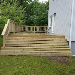 Nowo wybudowany drewniany taras przylegający do jasnoszarego domu z widocznymi schodami, balustradą i pojedynczym krzesłem na górnym poziomie, otoczony zielonym trawnikiem i drzewami.