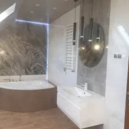 Nowoczesna łazienka na poddaszu z wanną narożną obudowaną brązowymi płytkami, białą szafką z umywalką, grzejnikiem drabinkowym i okrągłym lustrem pod trzema wiszącymi lampami.