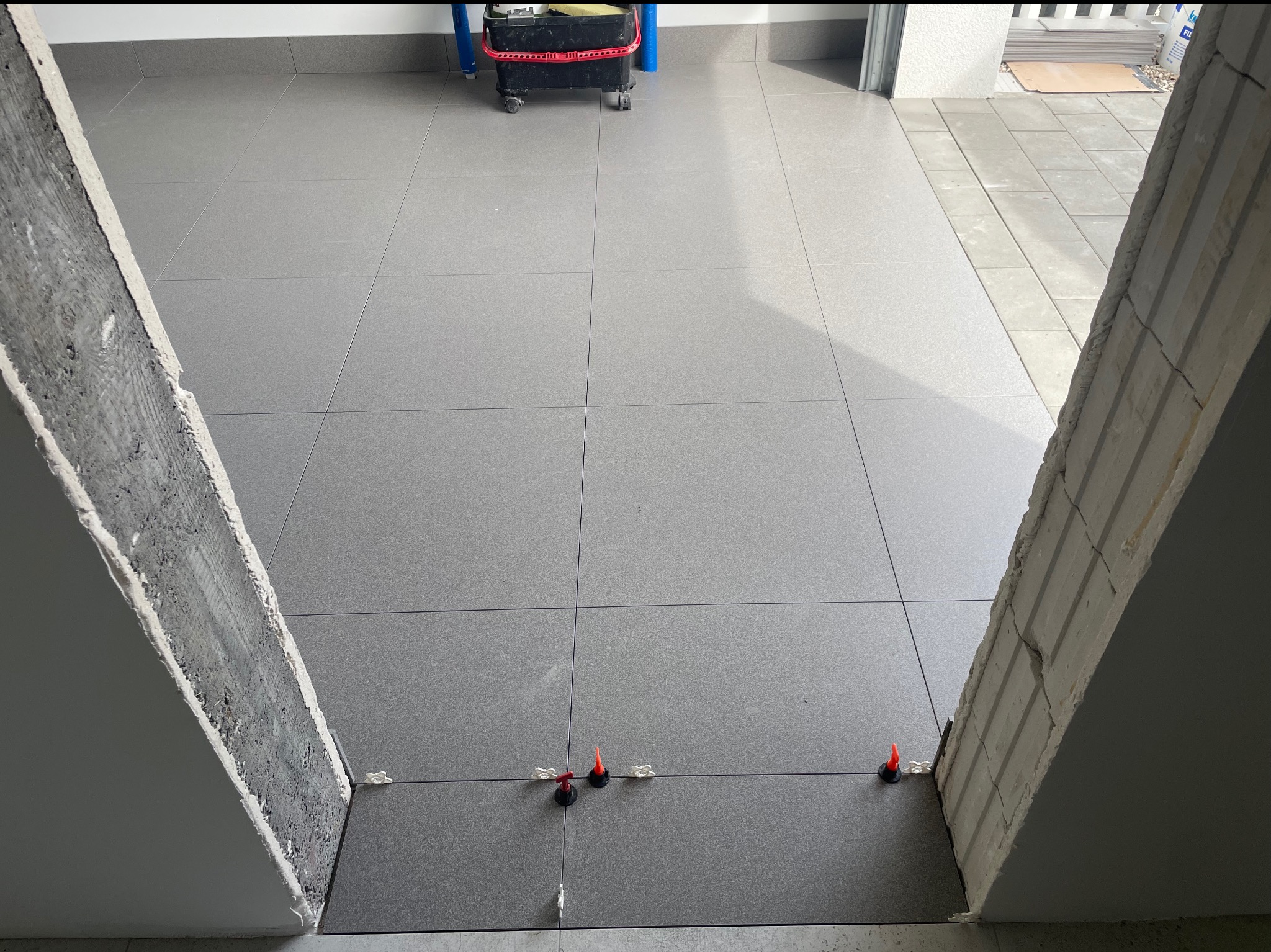 Szare płytki podłogowe ułożone w kwadraty, z systemem poziomowania płytek, widok z progu. W tle widoczne wiadro i inne materiały budowlane.
