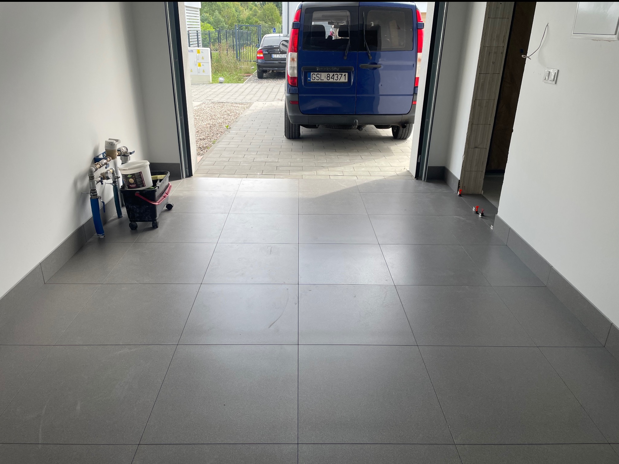 Szare płytki podłogowe w garażu, widok na zewnątrz z zaparkowanym niebieskim busem. Częściowo widoczne narzędzia glazurnicze.