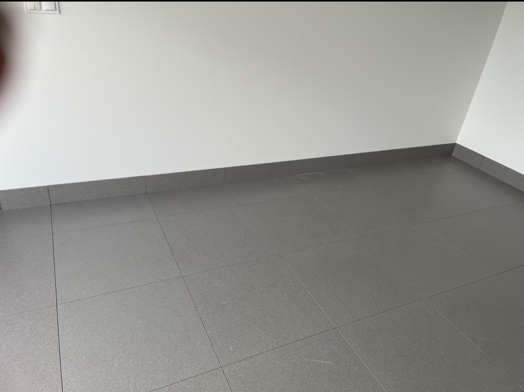 Szare, matowe płytki podłogowe ułożone w regularny wzór w pustym pokoju z białymi ścianami i szarą listwą przypodłogową. Widok z góry, neutralne oświetlenie.