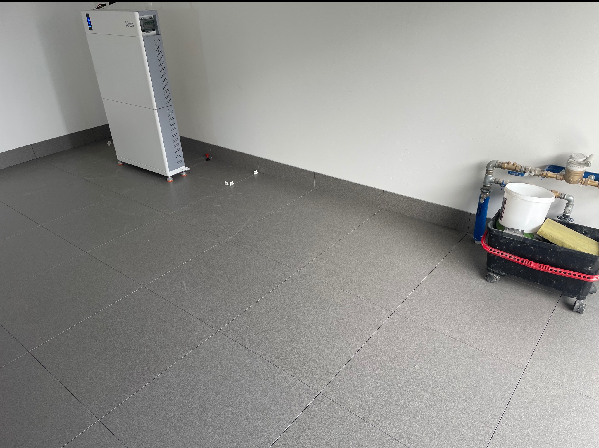 Wykończona szara podłoga z płytek w pomieszczeniu z białymi ścianami. Widoczne narzędzia glazurnicze i biała szafa. Minimalistyczny, nowoczesny design.