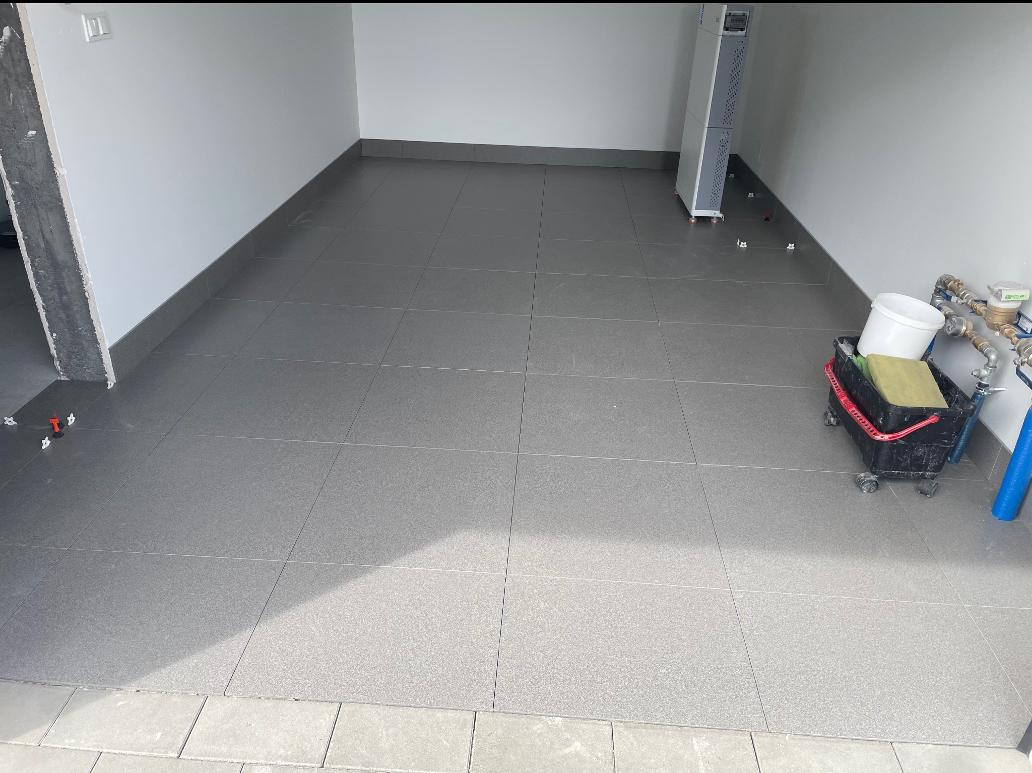 Szare płytki podłogowe ułożone w garażu, widoczne narzędzia glazurnicze i fragment instalacji. Minimalistyczny styl, świeżo wykończona podłoga z fugami.