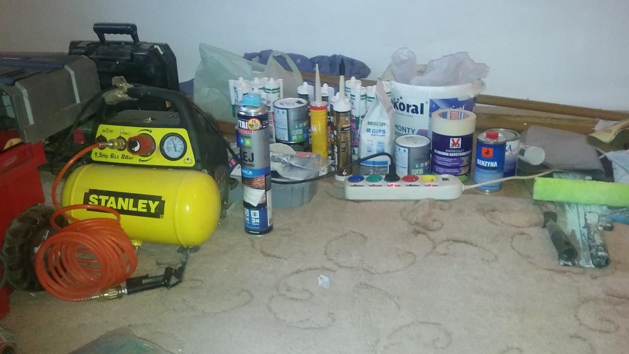 Stanowisko pracy z kompresorem Stanley, przedłużaczem, puszkami farb, gipsu i innymi materiałami budowlanymi na dywanie.