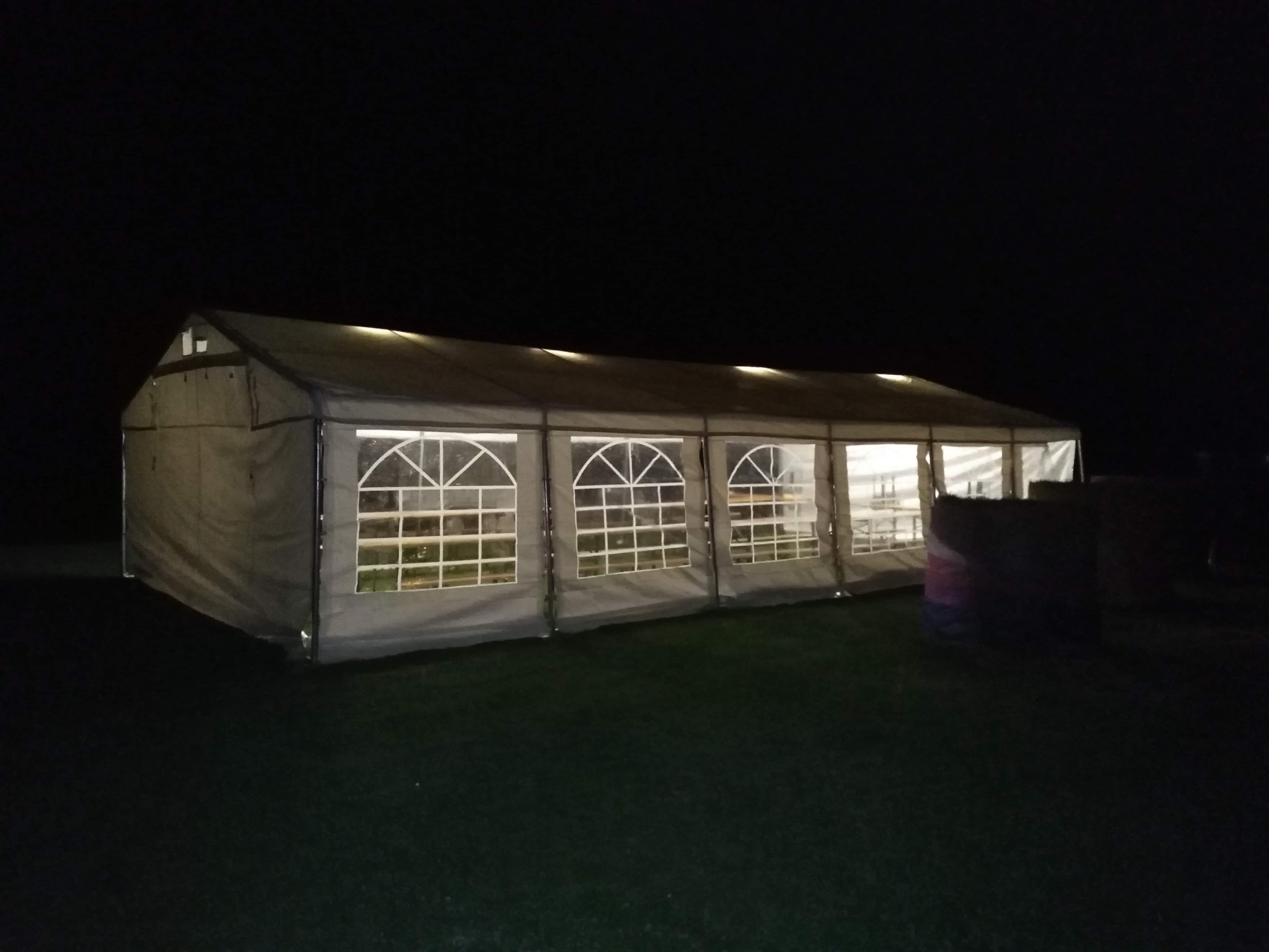 Rozświetlony namiot imprezowy z oknami, ustawiony na trawniku w ciemności nocy.