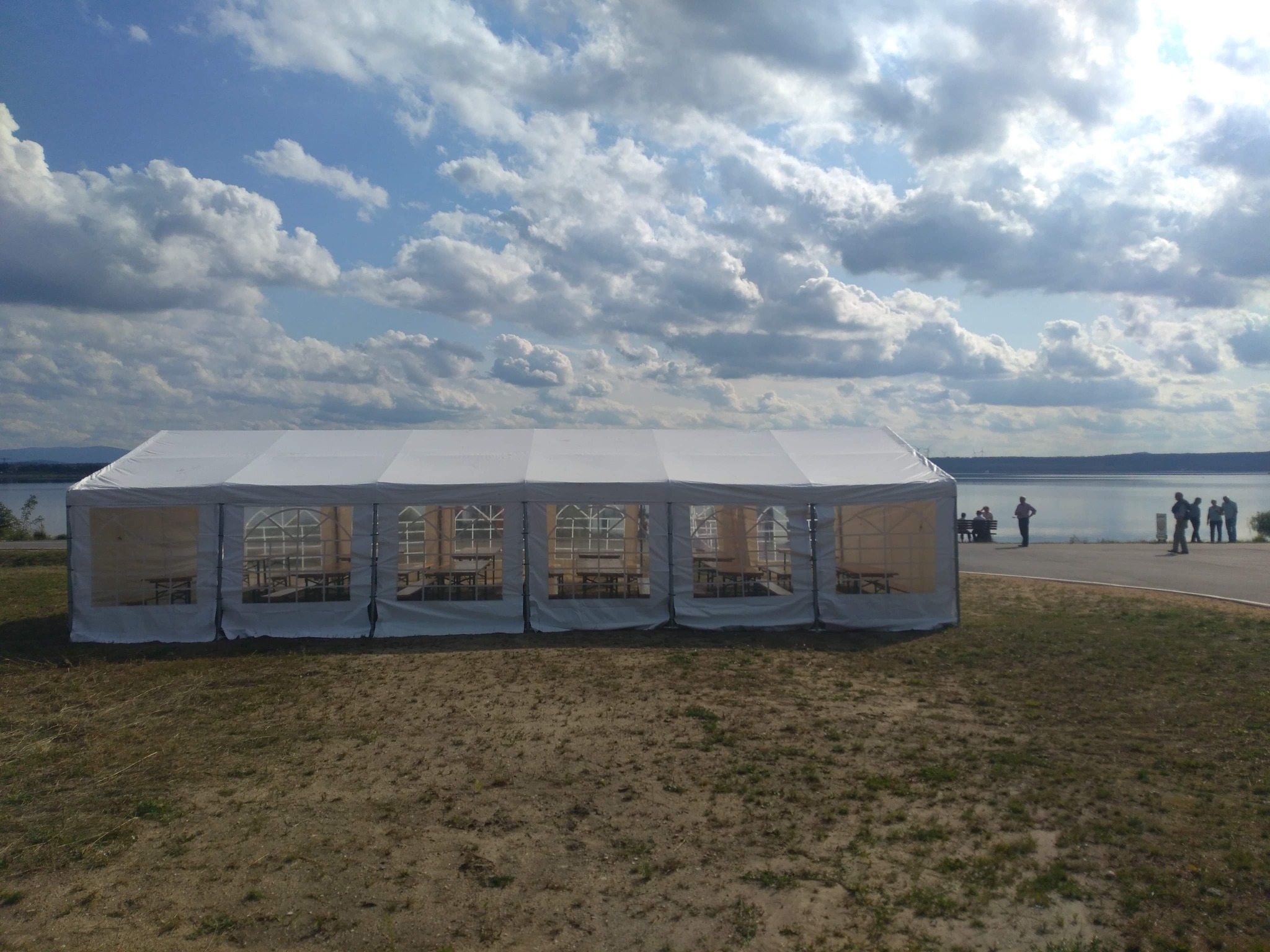 Biały namiot imprezowy z przezroczystymi ścianami bocznymi ustawiony na trawiastym terenie z widokiem na jezioro i grupę osób w oddali, pod błękitnym niebem z obłokami.