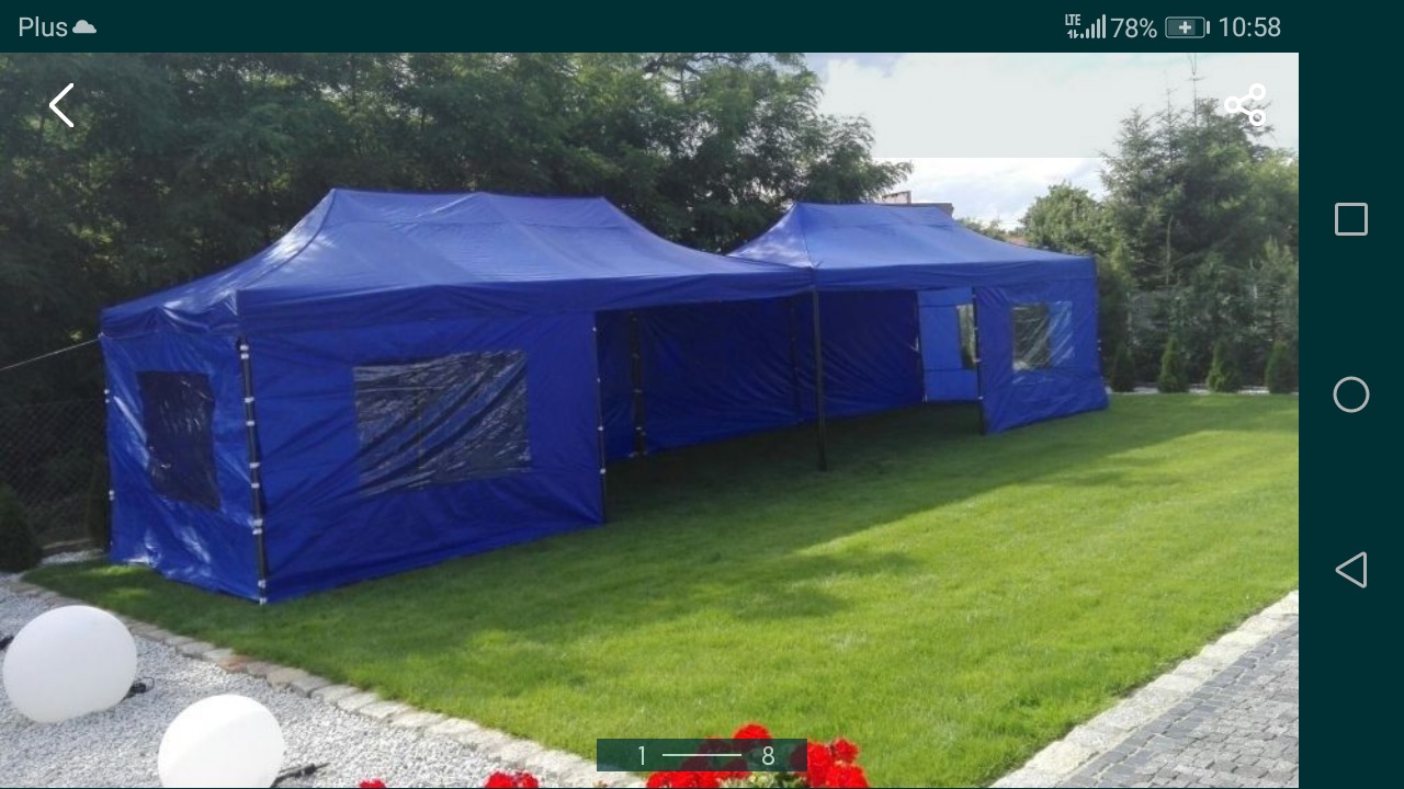 Dwa połączone, rozłożone namioty w kolorze niebieskim z oknami, ustawione na trawniku obok ścieżki z kostki brukowej i rabaty kwiatowej.