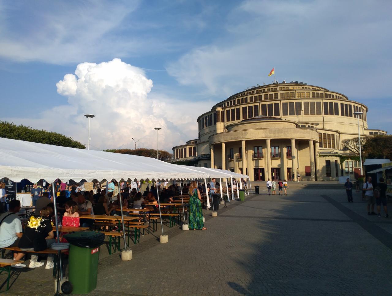 Biały namiot plenerowy z drewnianymi ławami i stolikami, ustawiony na tle Hali Stulecia we Wrocławiu, ludzie siedzący przy stolikach