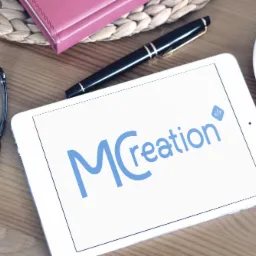 Tablet wyświetlający niebieskie logo 'MCreation' z ikoną kodu, obok czarny długopis, okulary, filiżanka kawy i różowy notatnik na drewnianym stole.