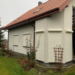 Zamknięte rolety zewnętrzne w kolorze kremowym na elewacji domu jednorodzinnego w Łazach, widoczne przycięte krzewy róż przed oknami.