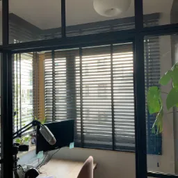 Biuro domowe z widokiem na budynek przez okno z ciemnymi żaluzjami poziomymi, częściowo zasłoniętymi przez czarną ramę szklanej ściany działowej.