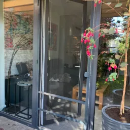 Otwarta, przesuwna moskitiera w metalowej ramie, widok z zewnątrz, z kwiatami fuksji w donicy po prawej stronie.