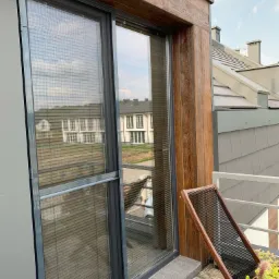 Szare okno przesuwne z wewnętrznymi listwami, drewniane wykończenie elewacji, balkon z drewnianą podłogą i minimalistycznym krzesłem.