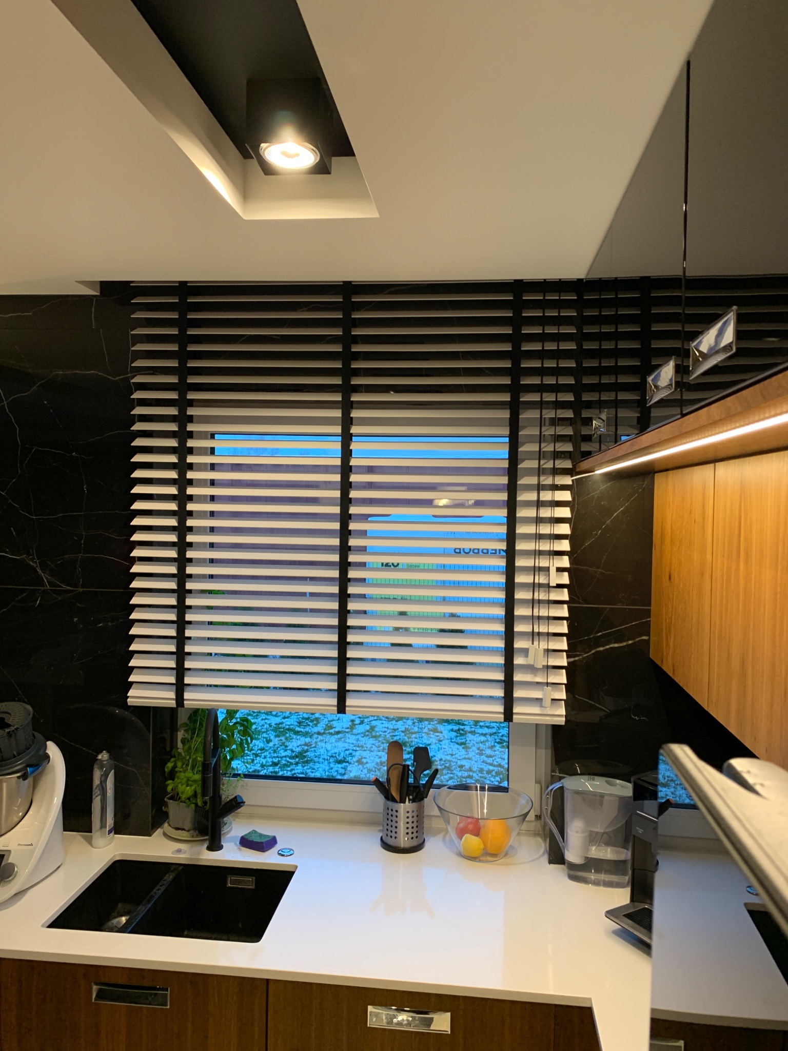 Kuchnia z czarnym marmurem na ścianie i drewnianymi szafkami, okno z poziomymi, białymi żaluzjami z czarnymi elementami, częściowo zasłonięte, w tle widok na zewnątrz.