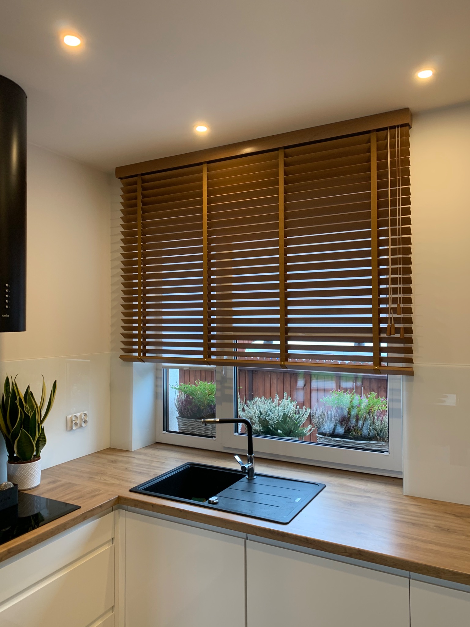 Kuchnia z drewnianymi żaluzjami w oknie, widok na ogród z roślinami w donicach.