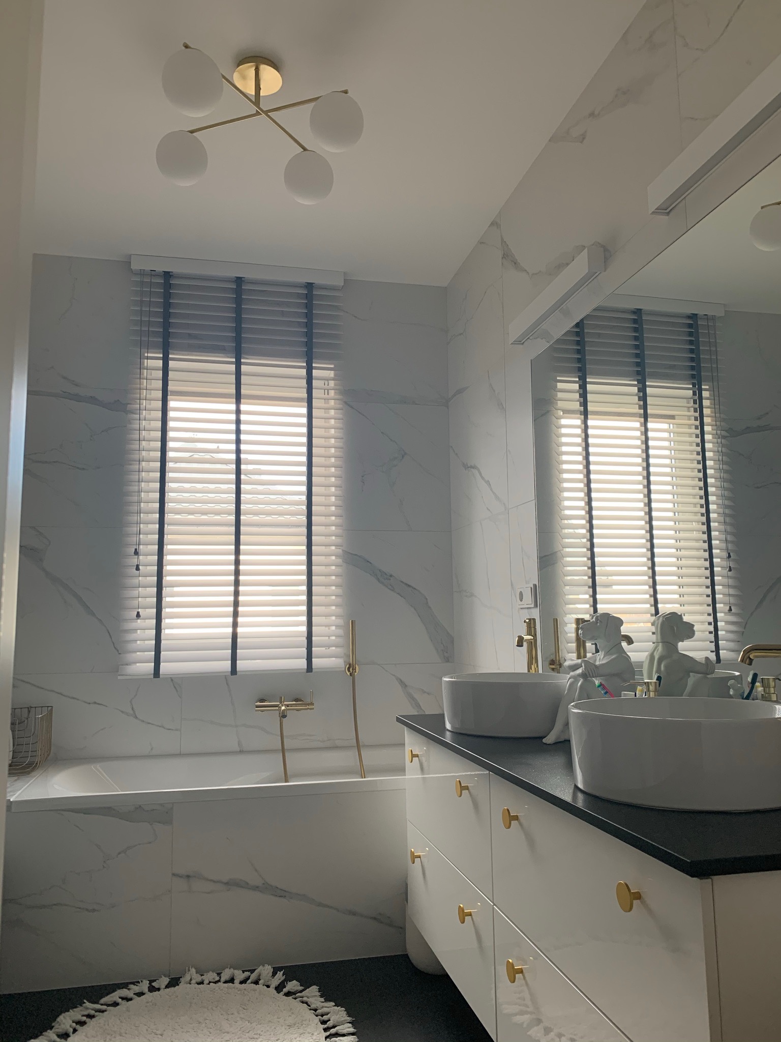 Nowoczesna łazienka z marmurowymi płytkami, białymi umywalkami nablatowymi, złotą armaturą i jasnymi żaluzjami drewnianymi z czarnymi akcentami na oknie.