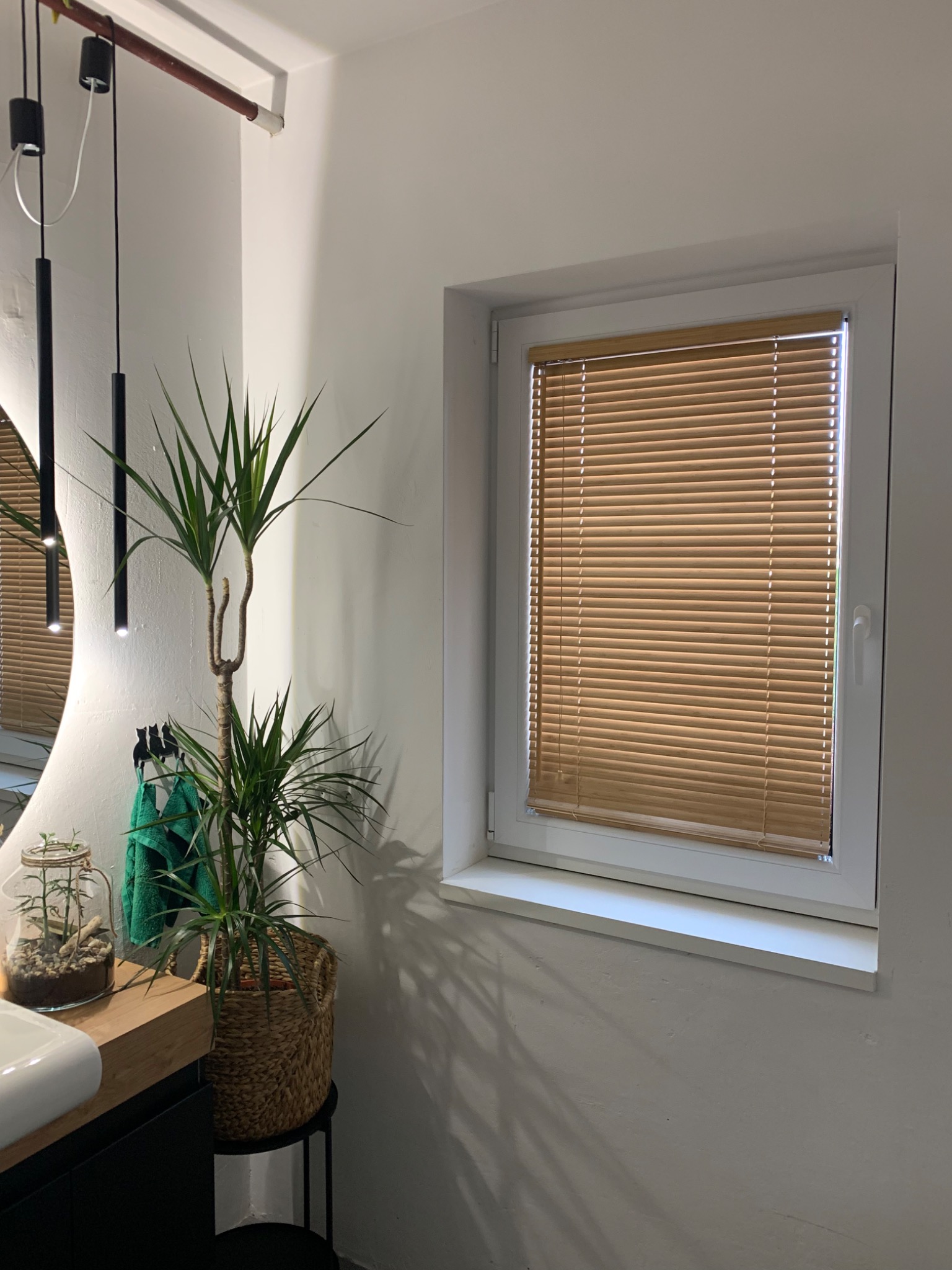 Jasne, minimalistyczne wnętrze łazienki z drewnianymi żaluzjami poziomymi w oknie, roślina doniczkowa w plecionym koszu i okrągłe lustro z podświetleniem.