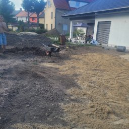 USŁUGI OGRODNICZE DAWID BANACH - Przygotowanie terenu pod trawnik: wyrównywanie ziemi i rozsypywanie piasku, widoczna taczka z ziemią, dom w tle.