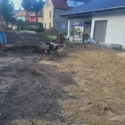 Przygotowanie terenu pod trawnik: wyrównywanie ziemi i rozsypywanie piasku, widoczna taczka z ziemią, dom w tle.