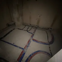 Surowe wnętrze w trakcie prac instalacyjnych: rury grzewcze (czerwone i niebieskie) ułożone na podłodze, widoczne połączenia i mocowania, ściany w stanie surowym, słabe oświetlenie.