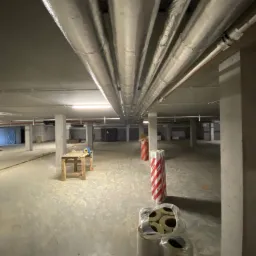 Montaż izolowanych rur wentylacyjnych w surowym pomieszczeniu, widoczne materiały izolacyjne w folii i prowizoryczny stół roboczy z narzędziami.