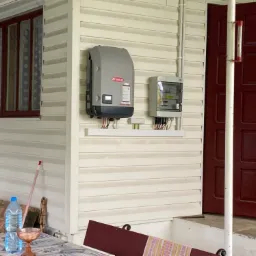 Szary inwerter Fronius zamontowany na elewacji domu obok skrzynki elektrycznej, z widocznymi kablami i elementami instalacji, na tle okna z ciemną ramą i czerwonych drzwi wejściowych.