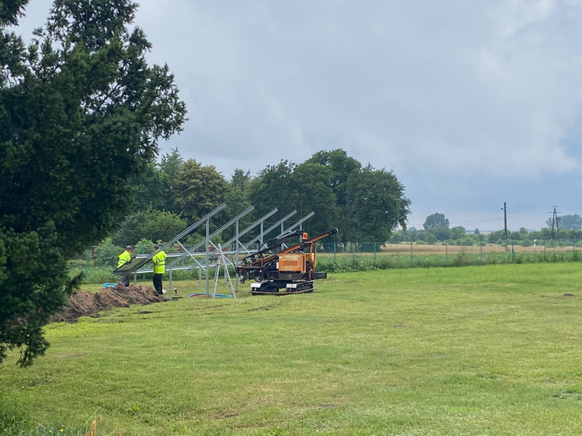 Montaż konstrukcji wsporczej pod panele słoneczne na zielonym terenie; w tle widoczny specjalistyczny sprzęt do wiercenia otworów pod fundamenty.