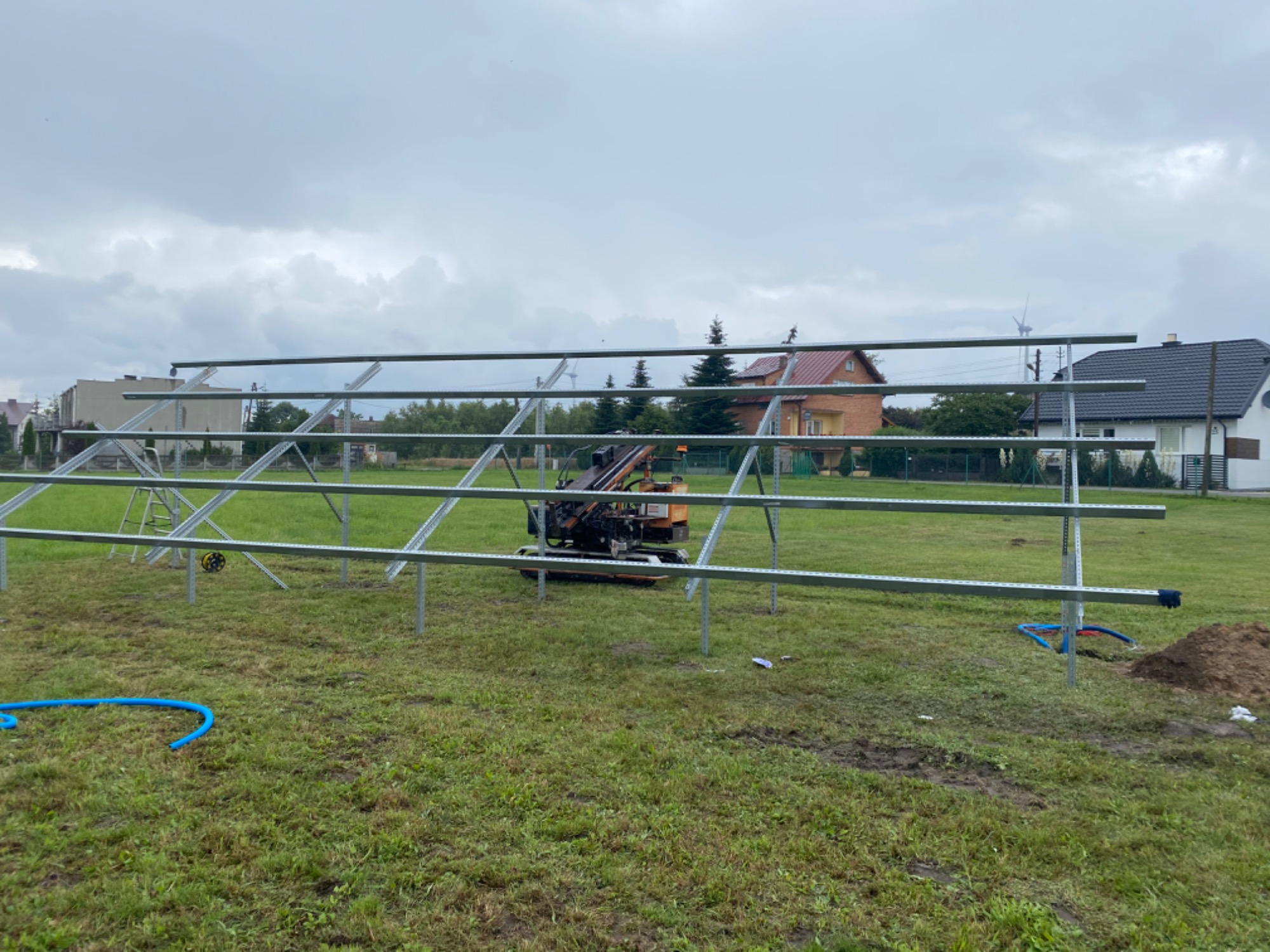 Montaż konstrukcji wsporczej pod panele słoneczne na trawiastym terenie, w tle domy jednorodzinne i pochmurne niebo. Widoczna wiertnica do gruntu.