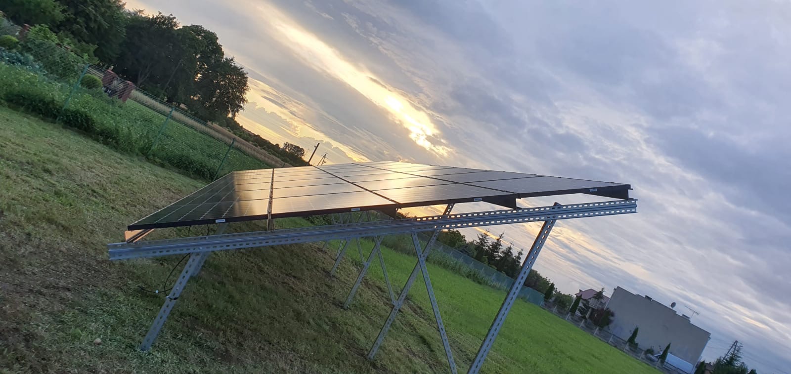 Instalacja paneli słonecznych na metalowej konstrukcji na trawiastym terenie, w tle zachodzące słońce i pochmurne niebo.