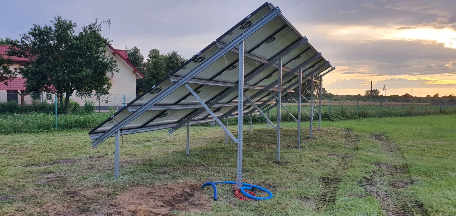 Montaż paneli słonecznych na konstrukcji wsporczej, widok od spodu, z widocznymi przewodami i elementami instalacji, na tle wiejskiego krajobrazu o zmierzchu.