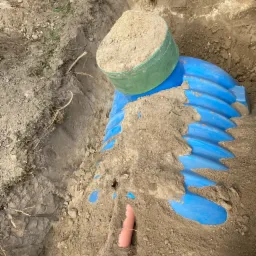 Niebieski element drenażowy w wykopie, częściowo zasypany piaskiem, z zielonym pojemnikiem na górze, widoczna rura kanalizacyjna.