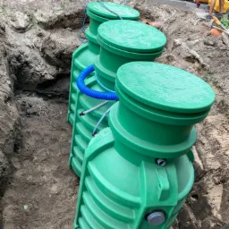 Trzy zielone zbiorniki połączone niebieskimi rurami, umieszczone w wykopie ziemnym, widoczne narzędzia i drewniane ogrodzenie w tle.