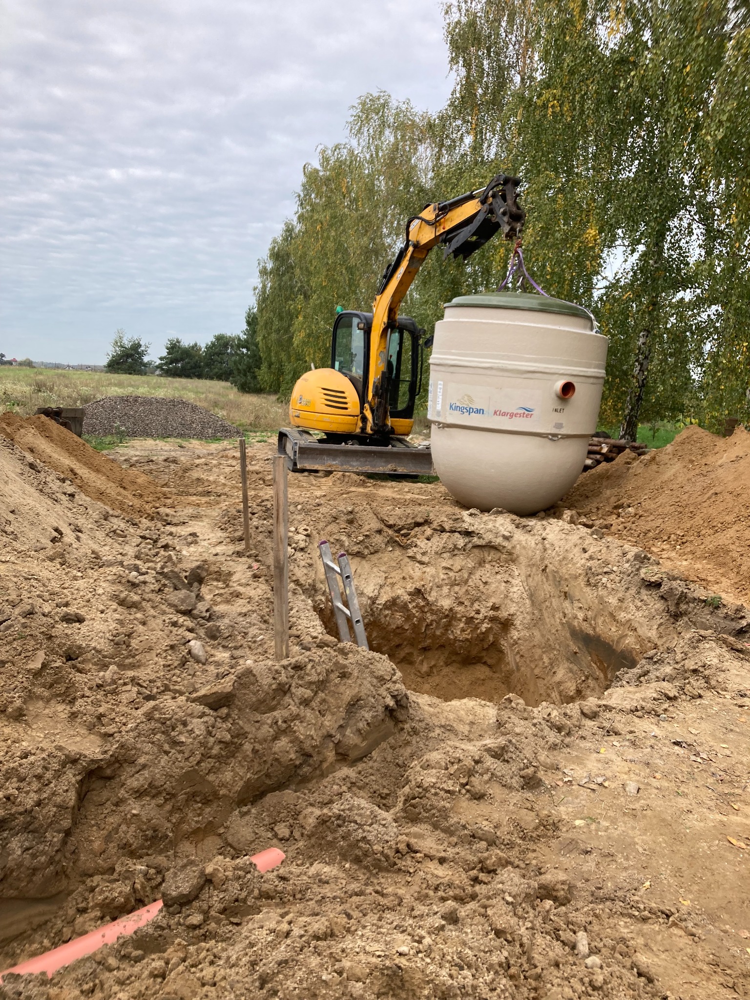 Żółta koparka umieszcza zbiornik z napisem Kingspan Klargester w wykopie z widoczną rurą kanalizacyjną w ziemi, na tle drzew i pochmurnego nieba.