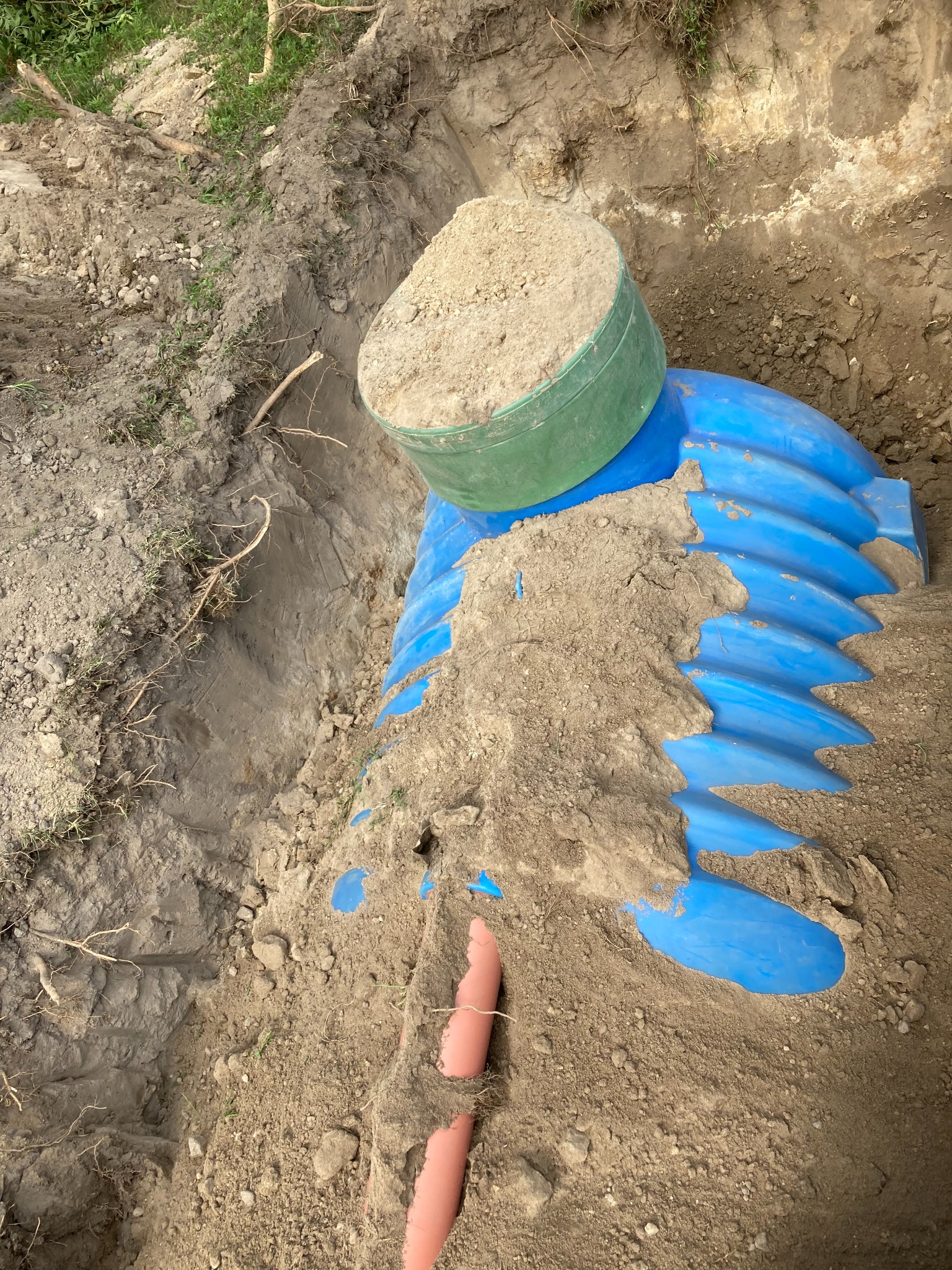 Niebieski element drenażowy w wykopie, częściowo zasypany piaskiem, z zielonym pojemnikiem na górze, widoczna rura kanalizacyjna.