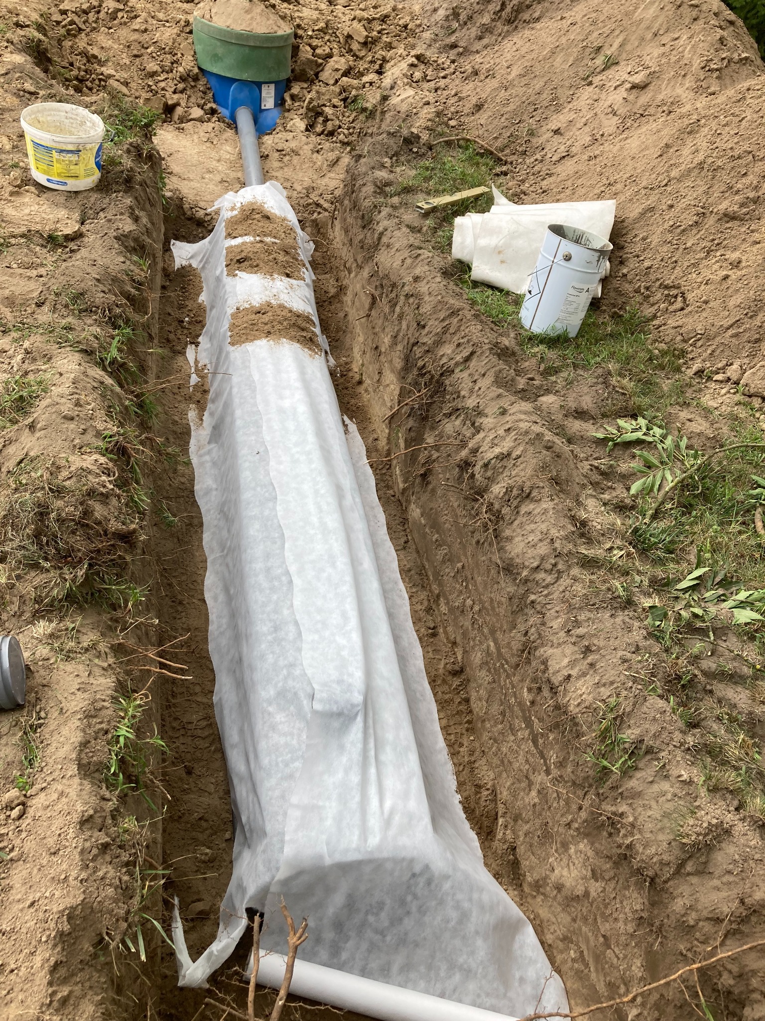 Wykop w ziemi z rurą owiniętą białą geowłókniną, częściowo zasypaną piaskiem, widoczny element instalacji i puszka farby obok wykopu.