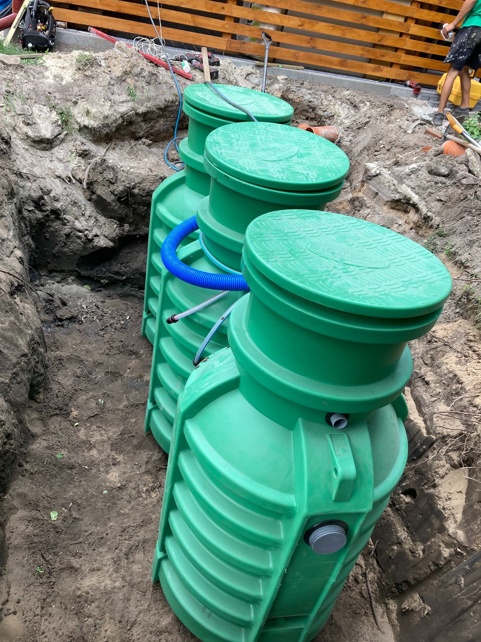 Trzy zielone zbiorniki połączone niebieskimi rurami, umieszczone w wykopie ziemnym, widoczne narzędzia i drewniane ogrodzenie w tle.