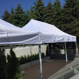 Biały namiot cateringowy z metalowym stelażem na drewnianym tarasie, w tle zielone drzewa iglaste i fragment szklanego dachu.